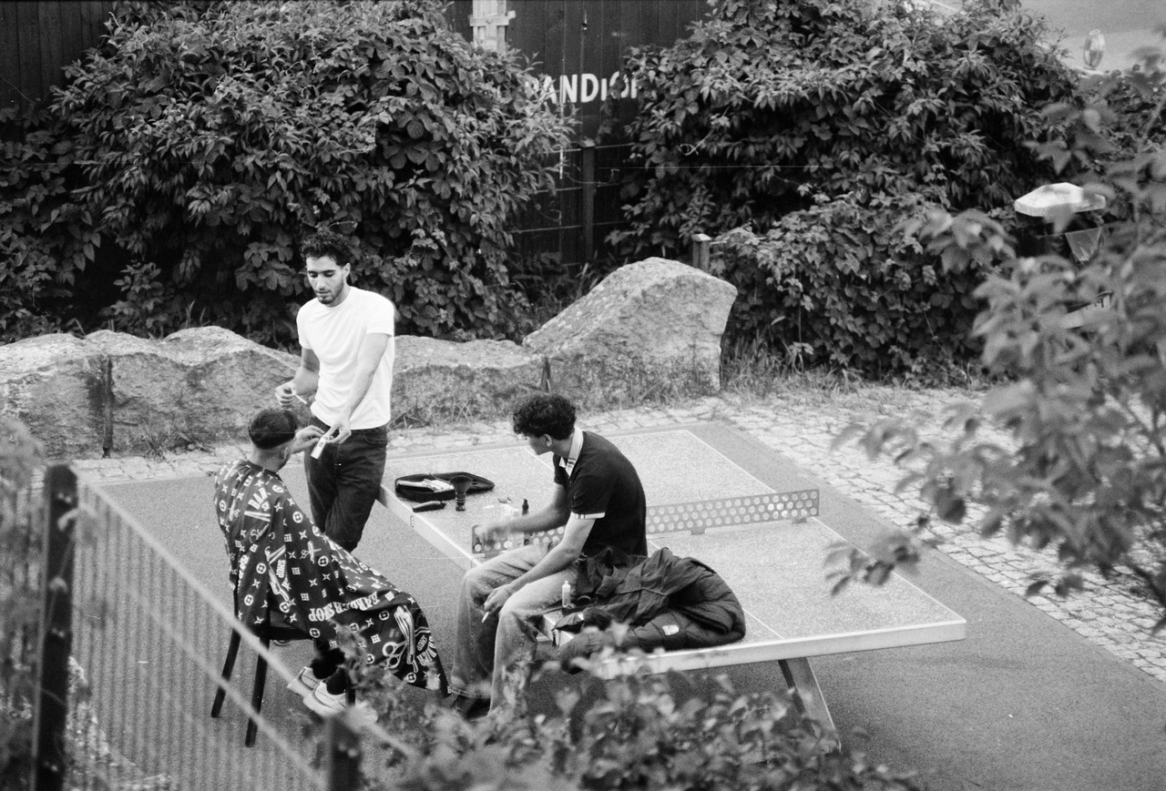 A man in a cape having his hair cut beside an outdoor table-tennis table, another man sitting at the table.