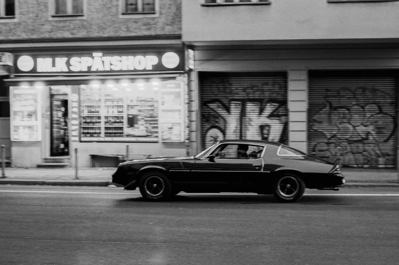 A dark muscle car driving past a lit corner shop and graffiti-covered shutters.