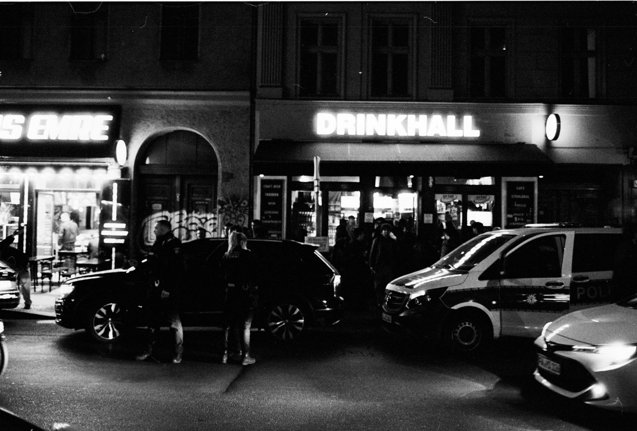 A police van and cars outside a lit late-night shop called Drinkhall.