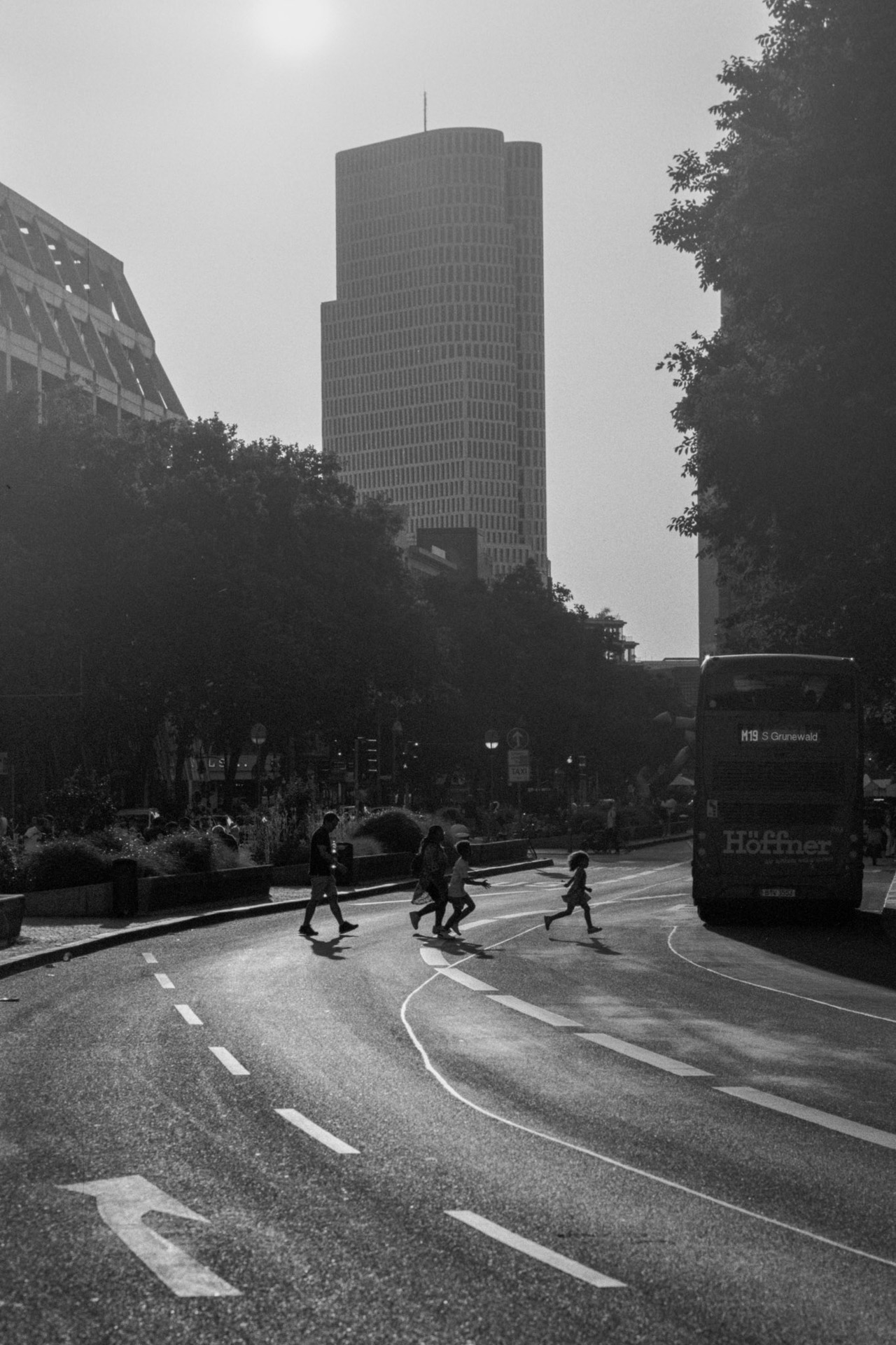 A family crossing a wide road toward a double-decker bus, a tower block behind.