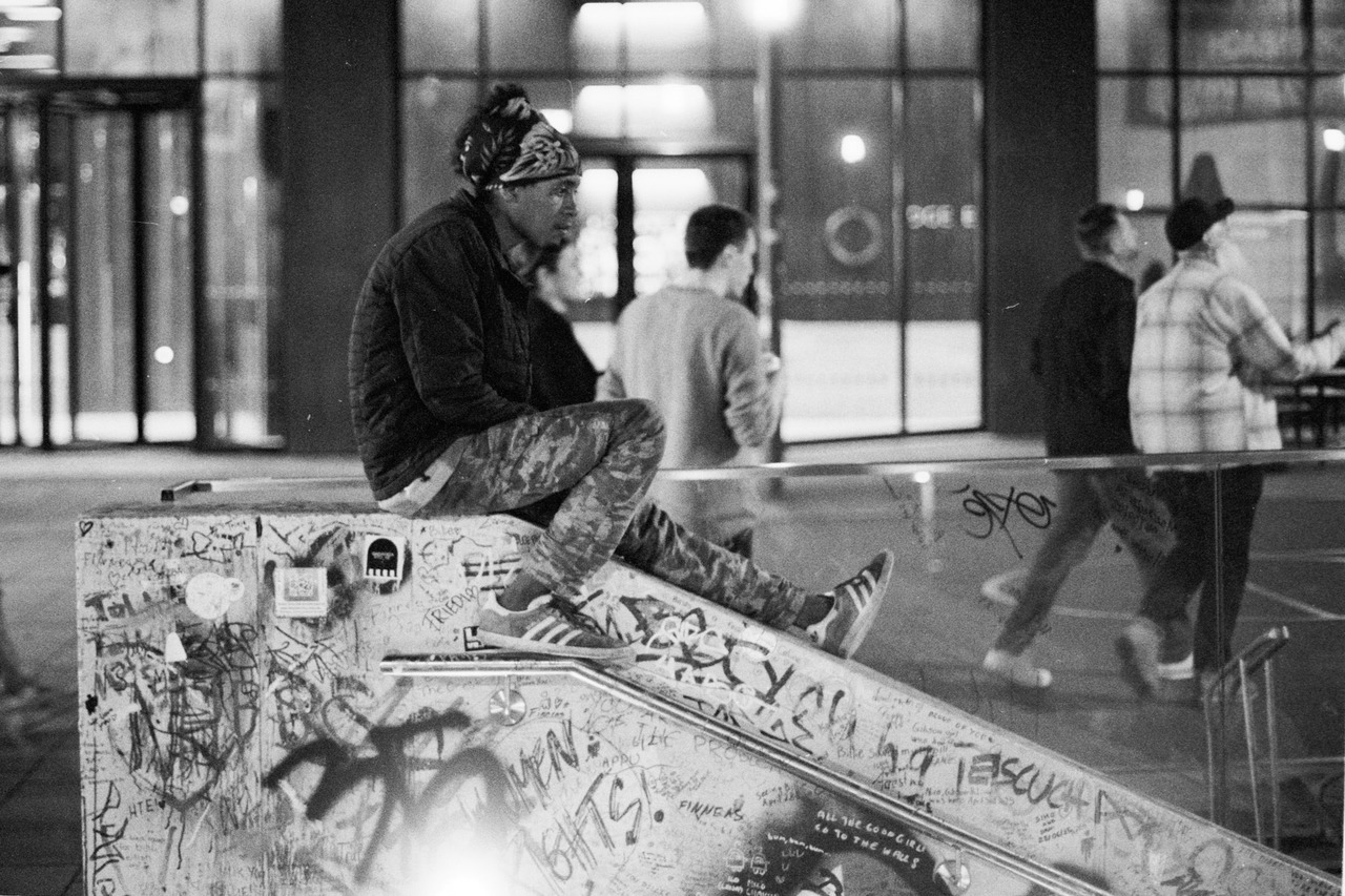 A man in camouflage trousers sitting on a graffiti-covered ledge at night, people walking behind.