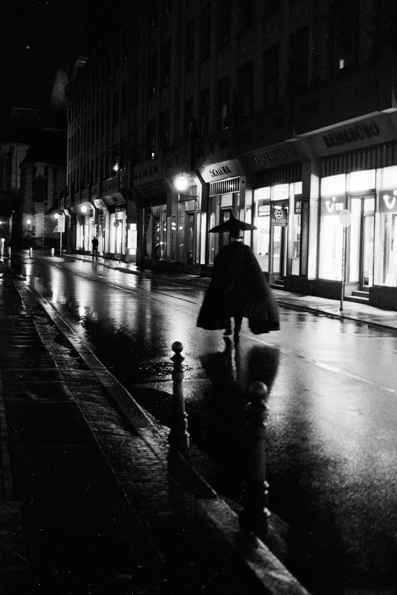 A person with an umbrella and a long coat walking down a wet street at night.