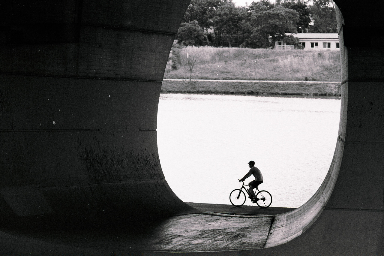A cyclist silhouetted in the rounded opening beneath a bridge, water behind.