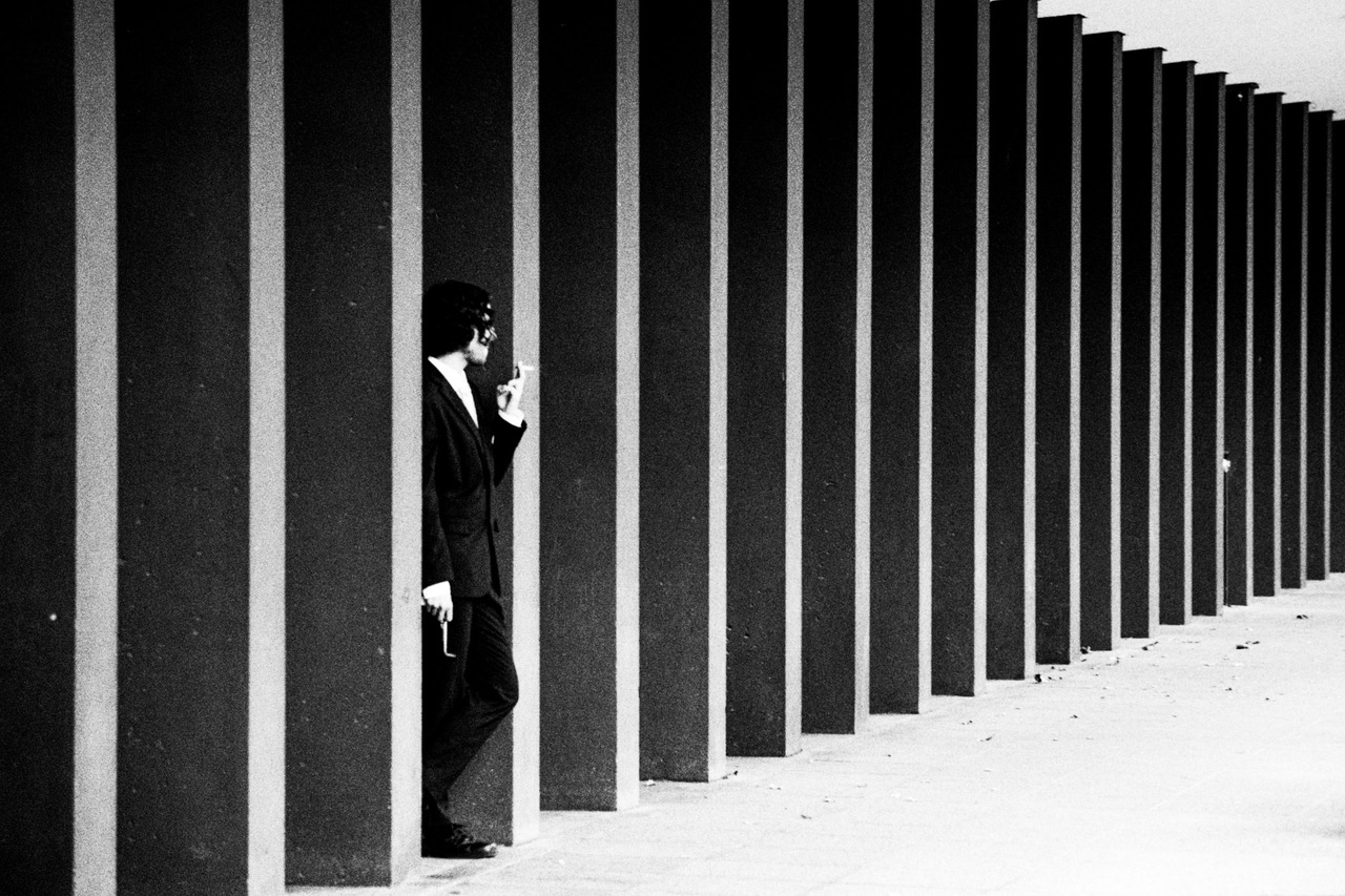 A man in a suit smoking, leaning against one of a long row of dark pillars.