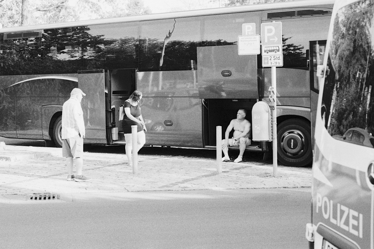 A shirtless man sitting in the open luggage hold of a coach while two people stand nearby; a police van in the foreground.