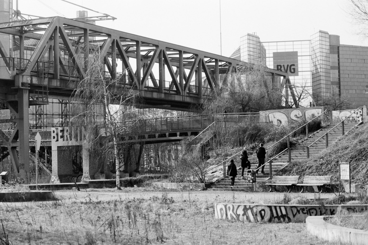 A steel truss railway bridge over a scrubby park, people climbing graffiti-lined steps below.