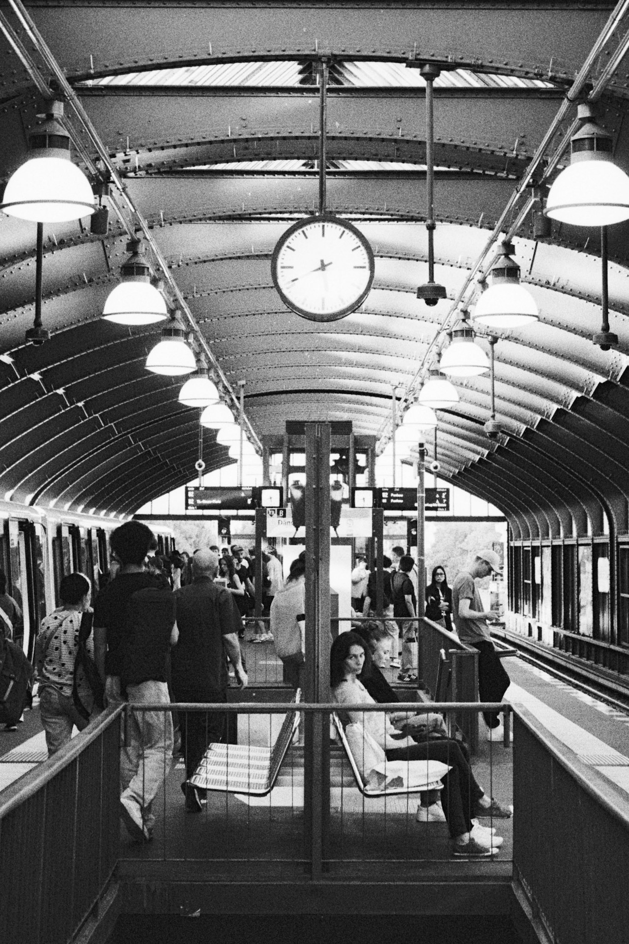 An elevated U-Bahn platform under an arched steel roof with a hanging clock, people waiting and walking.
