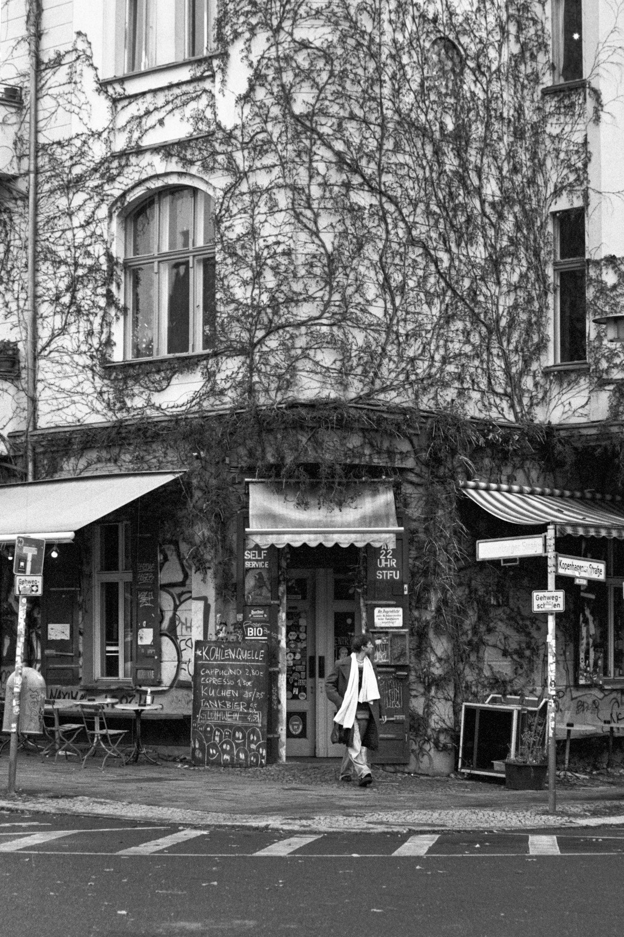 A man in a white scarf walking past a café on the ground floor of an ivy-covered building.