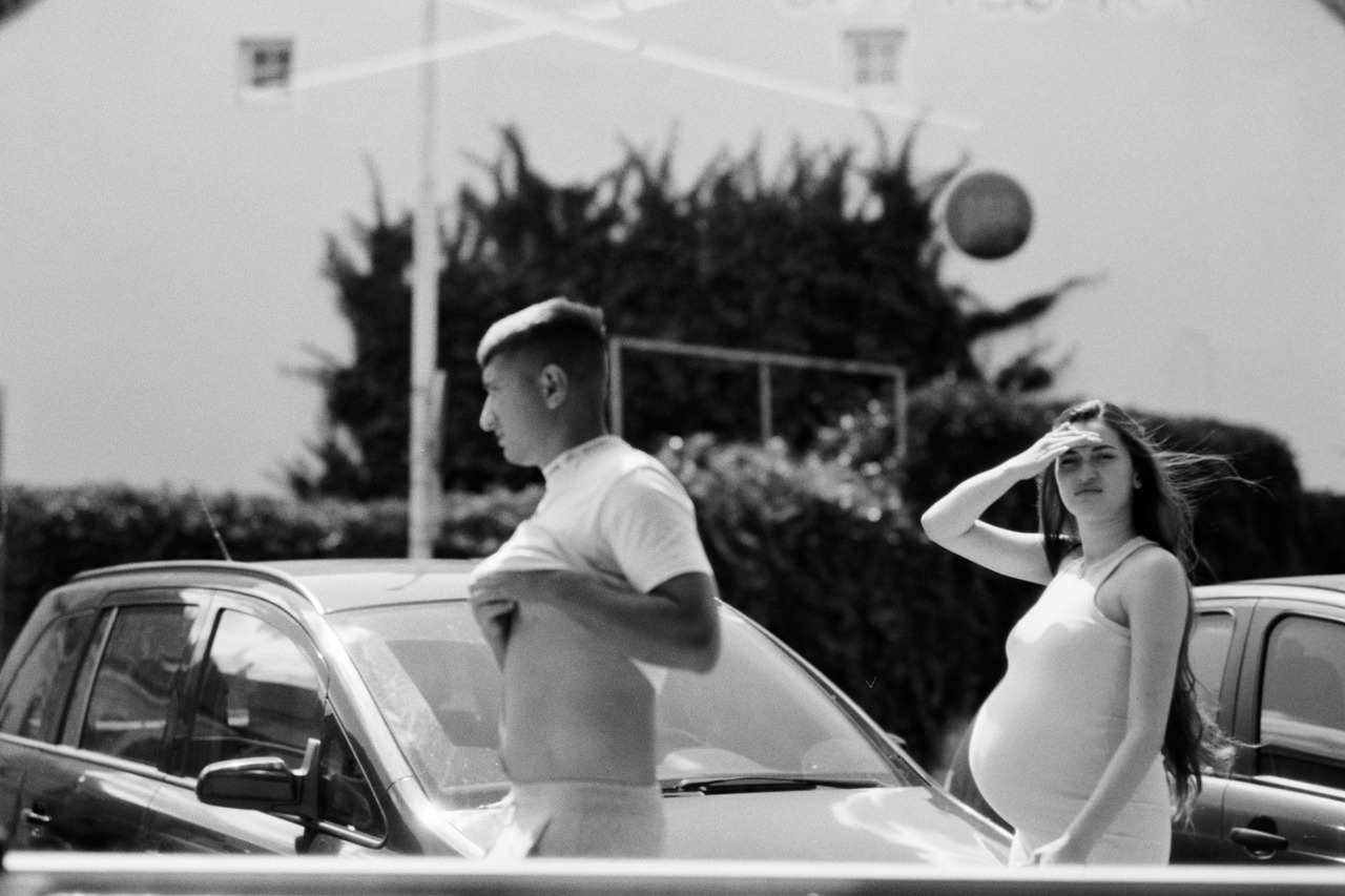 A young man and a pregnant woman shading her eyes, standing between parked cars.