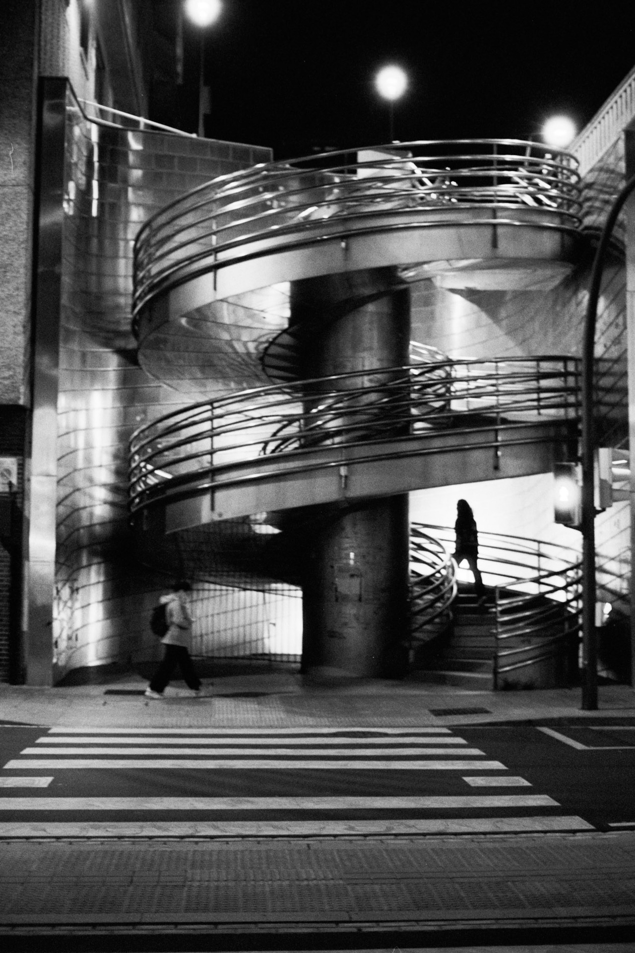A steel spiral staircase at a street corner at night, with two people passing.