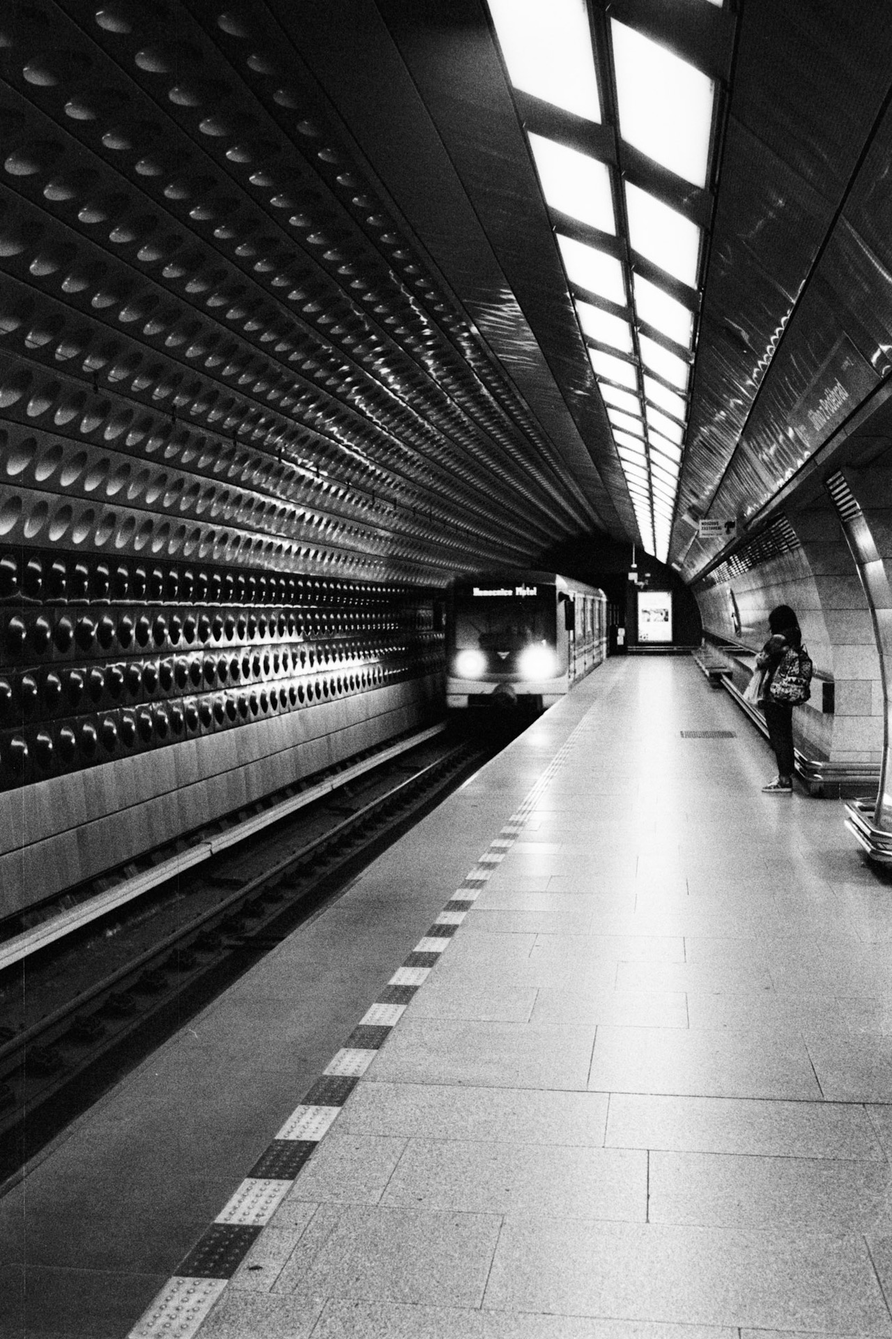 A train arriving at an underground platform with a studded curved wall, one woman waiting.