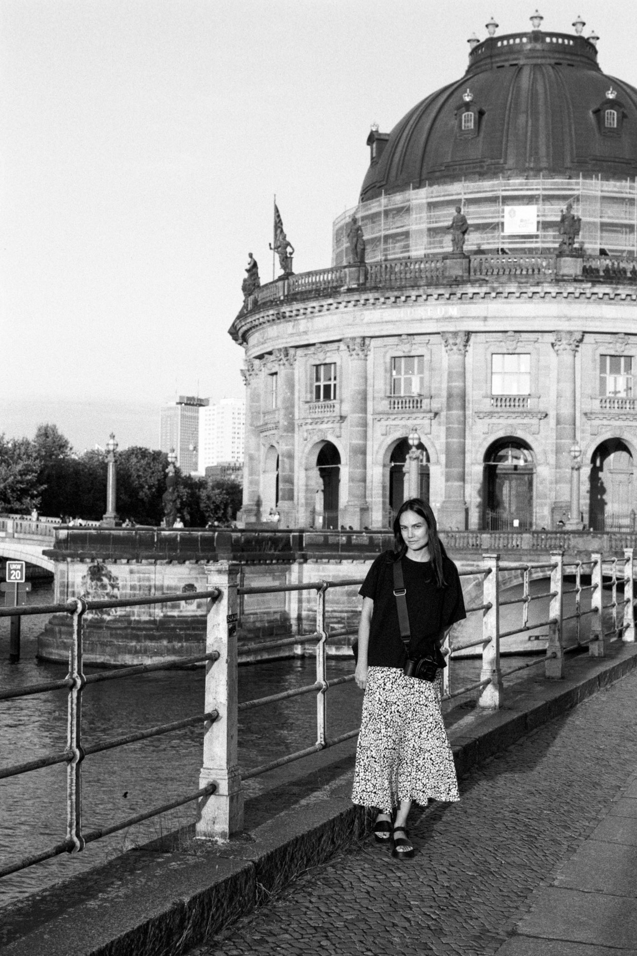 A woman in a black top and patterned skirt standing by a riverside railing, the Bode Museum behind.
