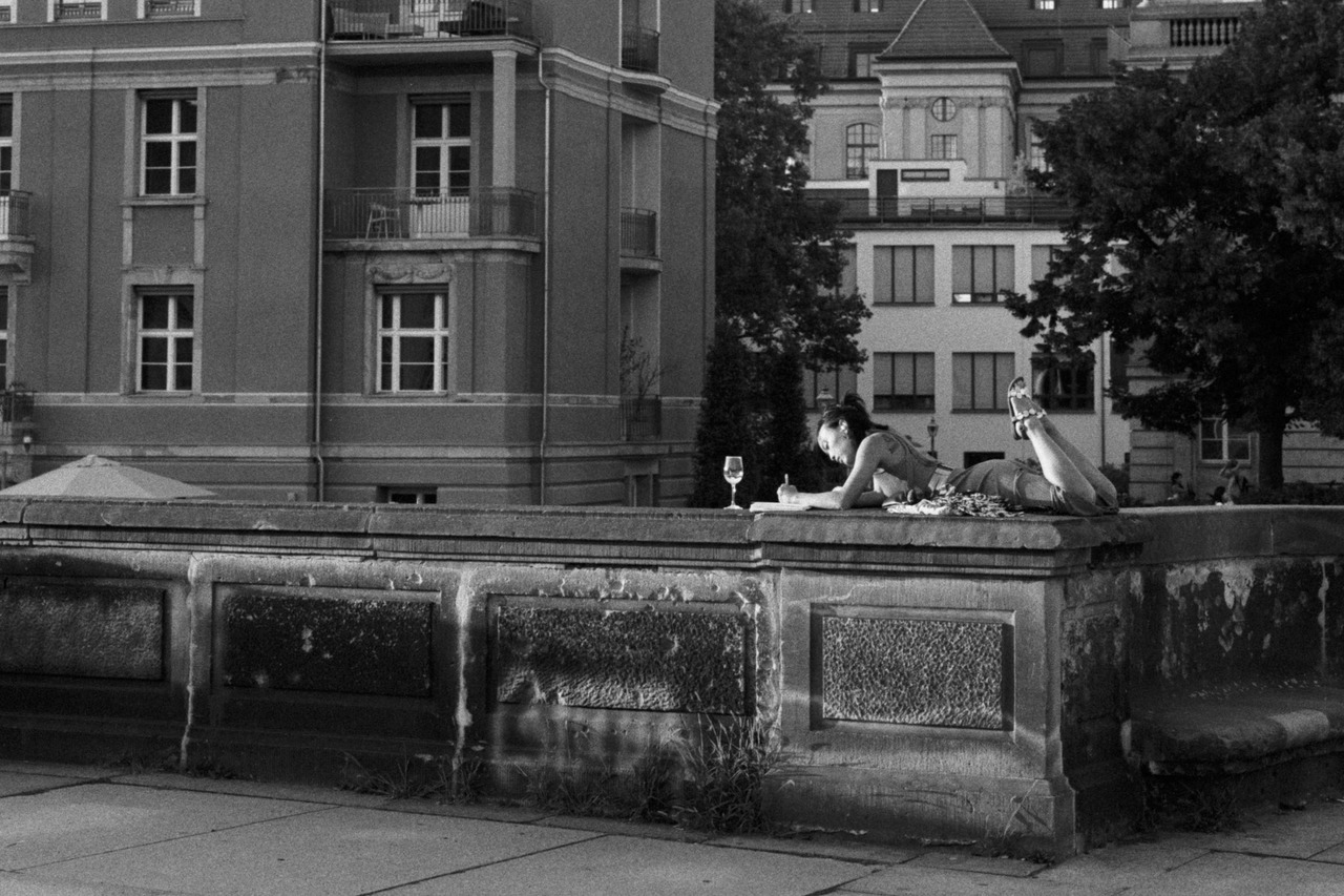 A woman lying on a stone wall, writing in a notebook next to a glass of wine.