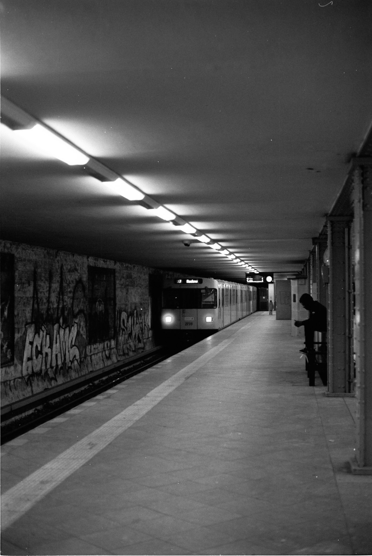 An underground platform with graffiti on the far wall and a train approaching, one person seated.