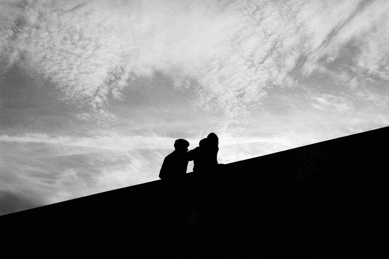 Two people silhouetted at the top of a dark slope against a cloudy sky.