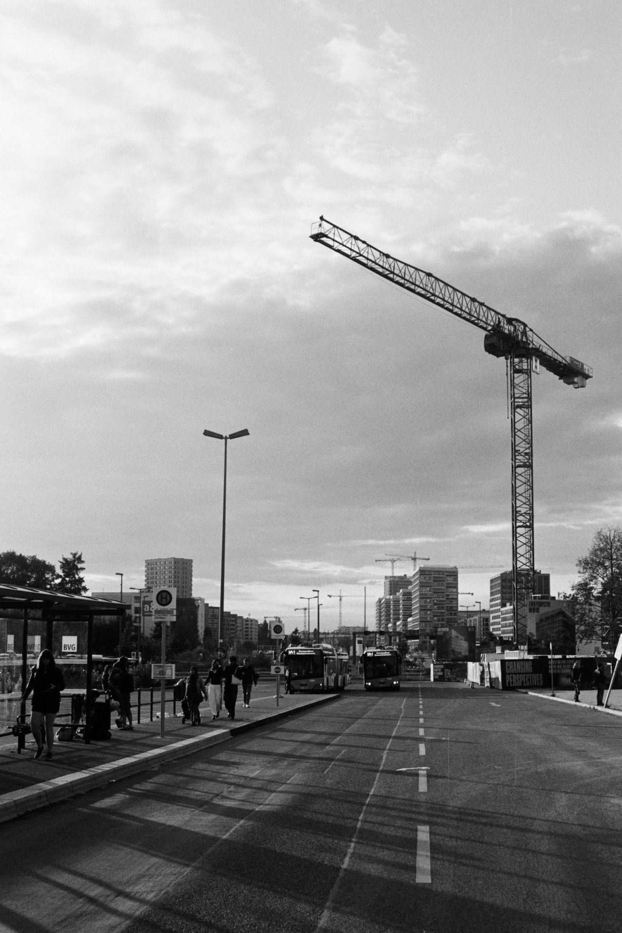 A tower crane above a bus stop, people waiting under a wide sky.