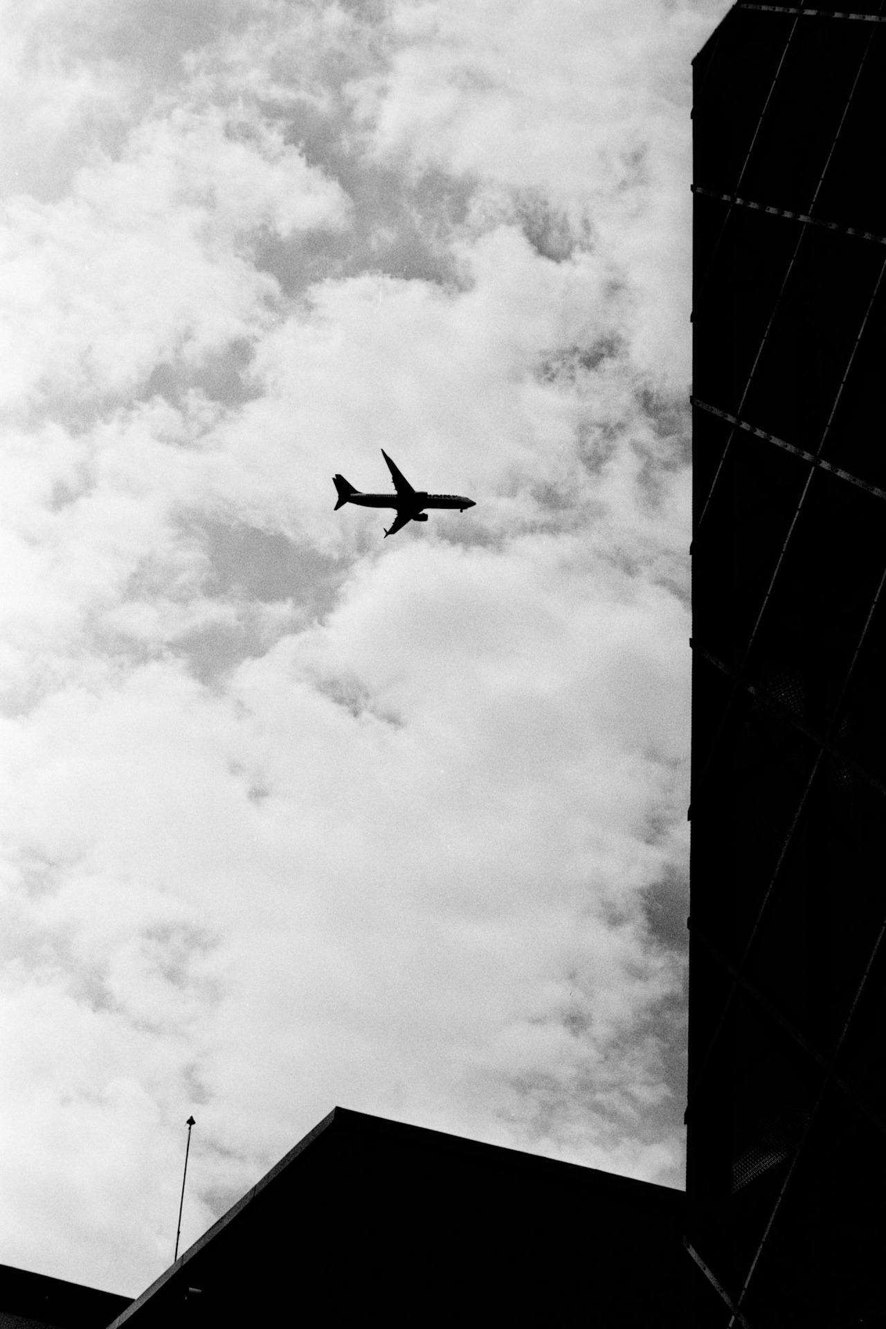 An airliner flying across a cloudy sky between the dark edges of buildings.