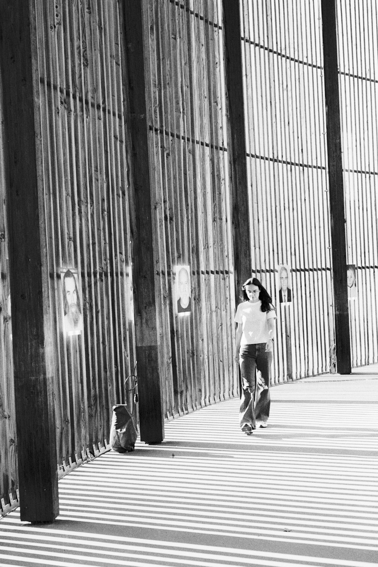 A woman walking through a curved wooden colonnade that casts striped shadows, portraits on the slats.