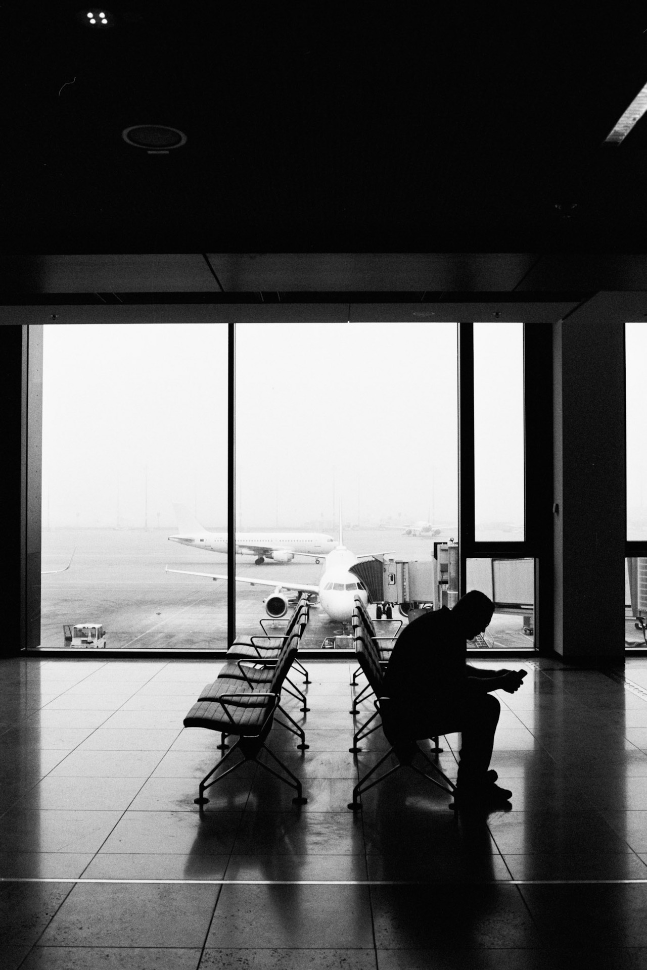 A seated silhouette in an airport lounge, planes on the apron beyond the windows.