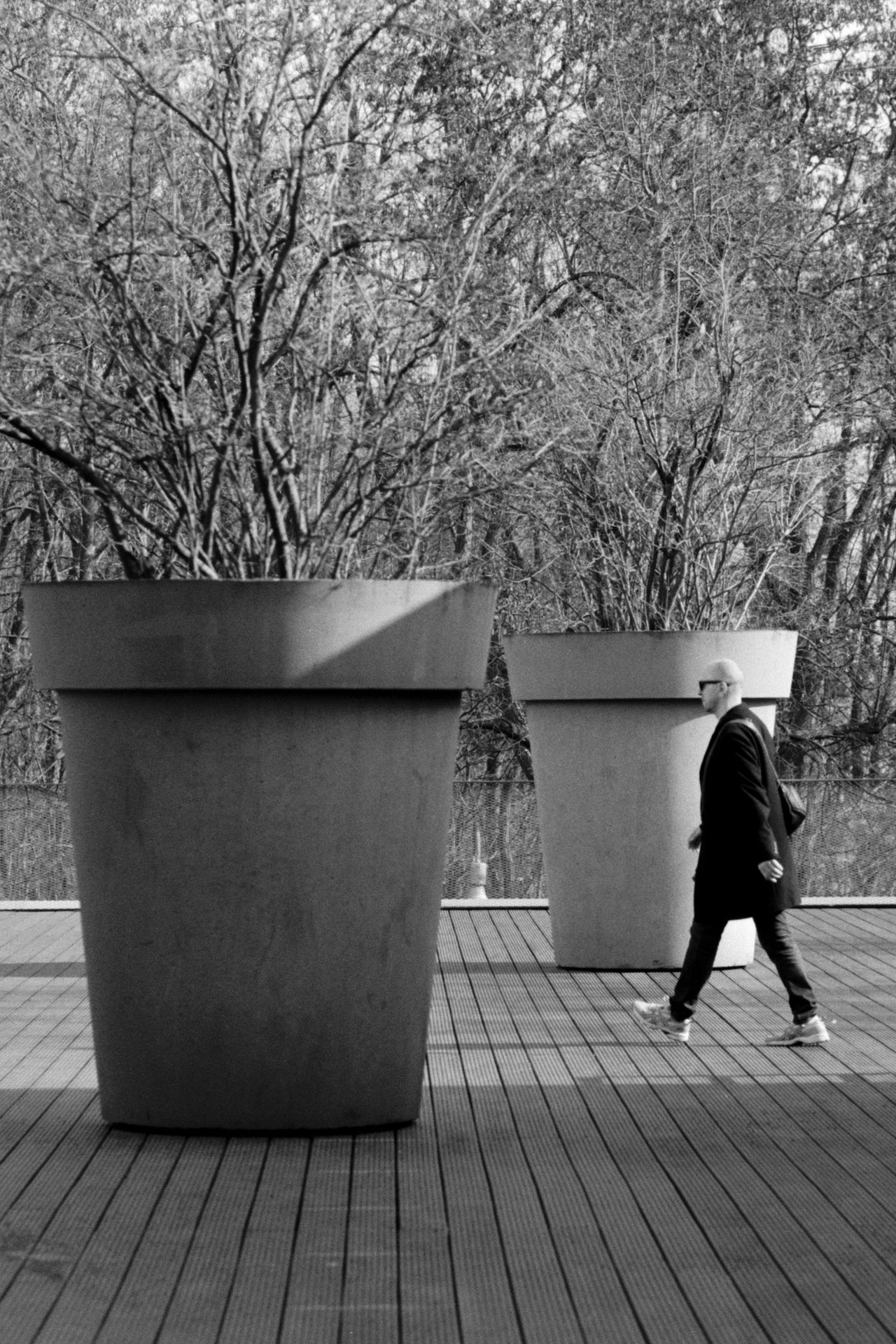 A man in a dark coat walking past two oversized concrete planters on a wooden deck.
