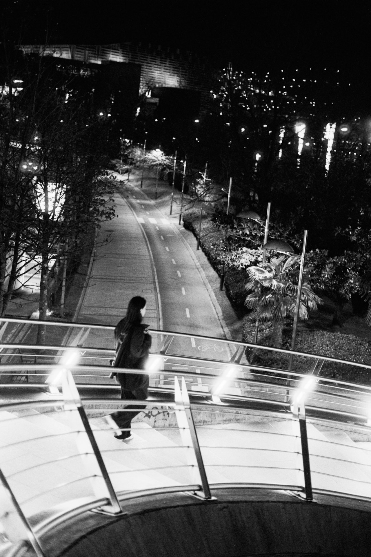 A woman crossing a curved walkway with lit railings above an empty road at night.
