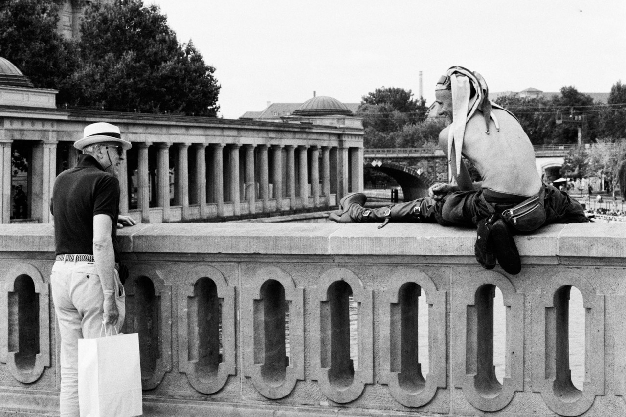 An older man in a hat and a shirtless man sitting on a stone bridge balustrade, a colonnade behind.