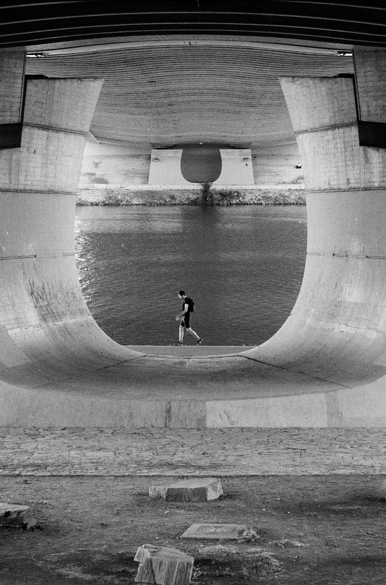 A man walking through the curved concrete underside of a bridge, the river visible beyond.