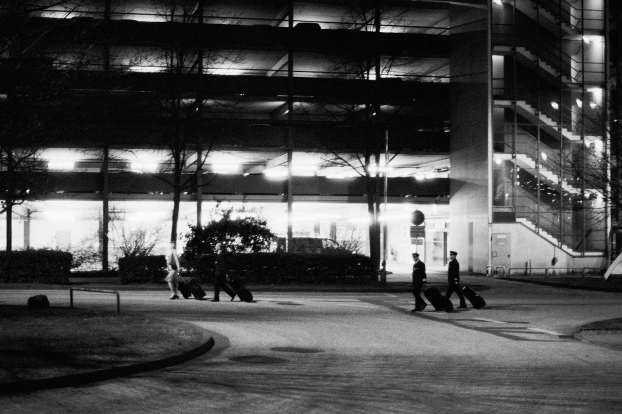 Airline crew pulling suitcases past a lit parking garage at night.