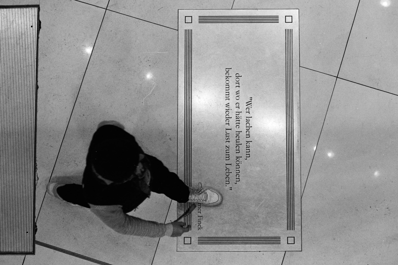Seen from above, a person standing on a floor plaque inscribed with a German quotation by Werner Finck.