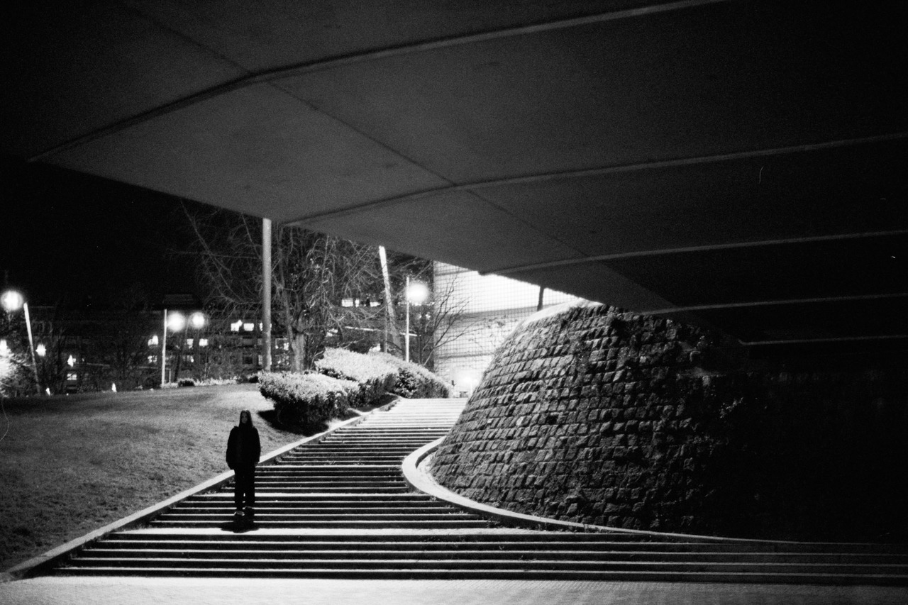 A person standing on wide steps under a concrete overhang at night, beside a stone wall.