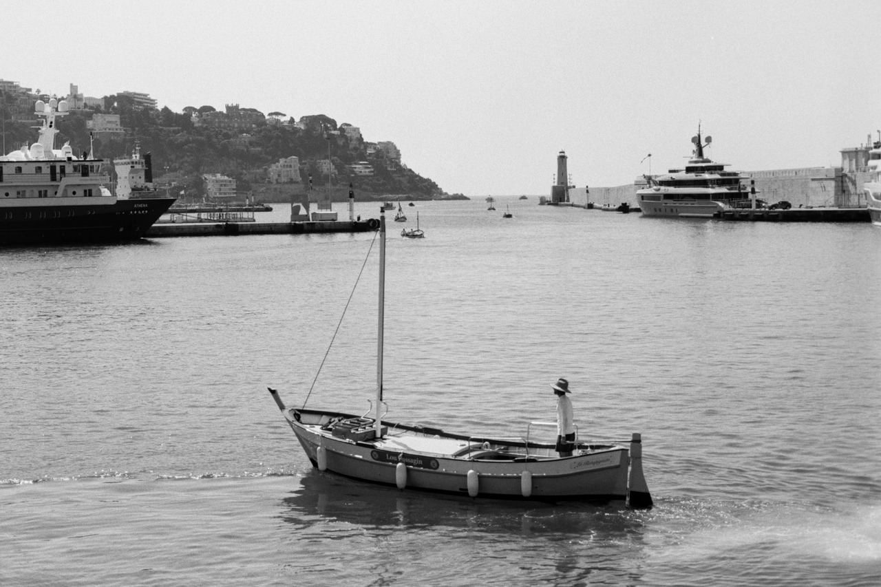 A man standing in a small wooden sailing boat in a harbour, large yachts moored behind.