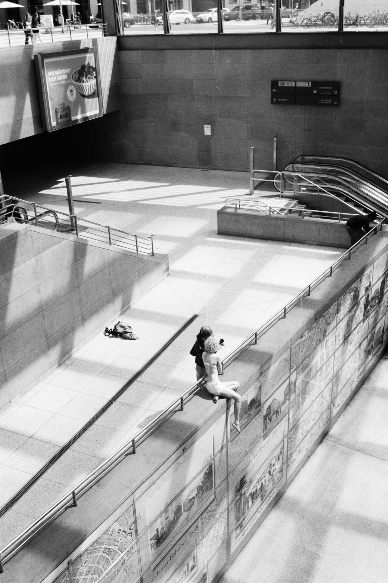 A station concourse seen from above, with a woman sitting on a sunlit ledge by the stairs.