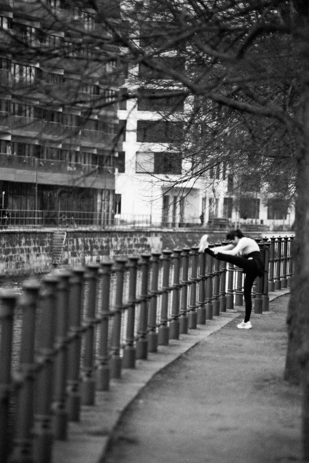 A woman stretching one leg along a riverside railing under bare trees.