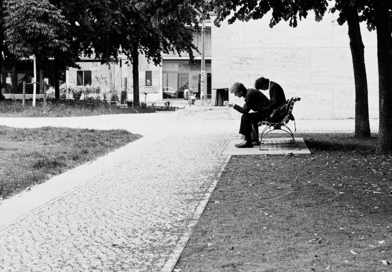 Two men in dark suits sitting on a park bench, one leaning forward over his phone.