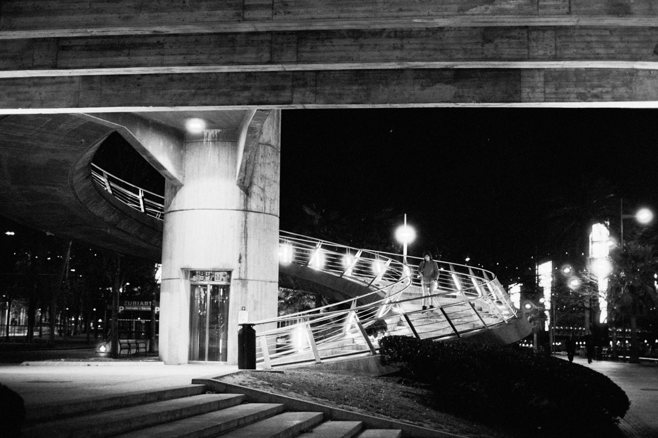 A figure standing on a lit spiral ramp beneath a concrete overpass at night.