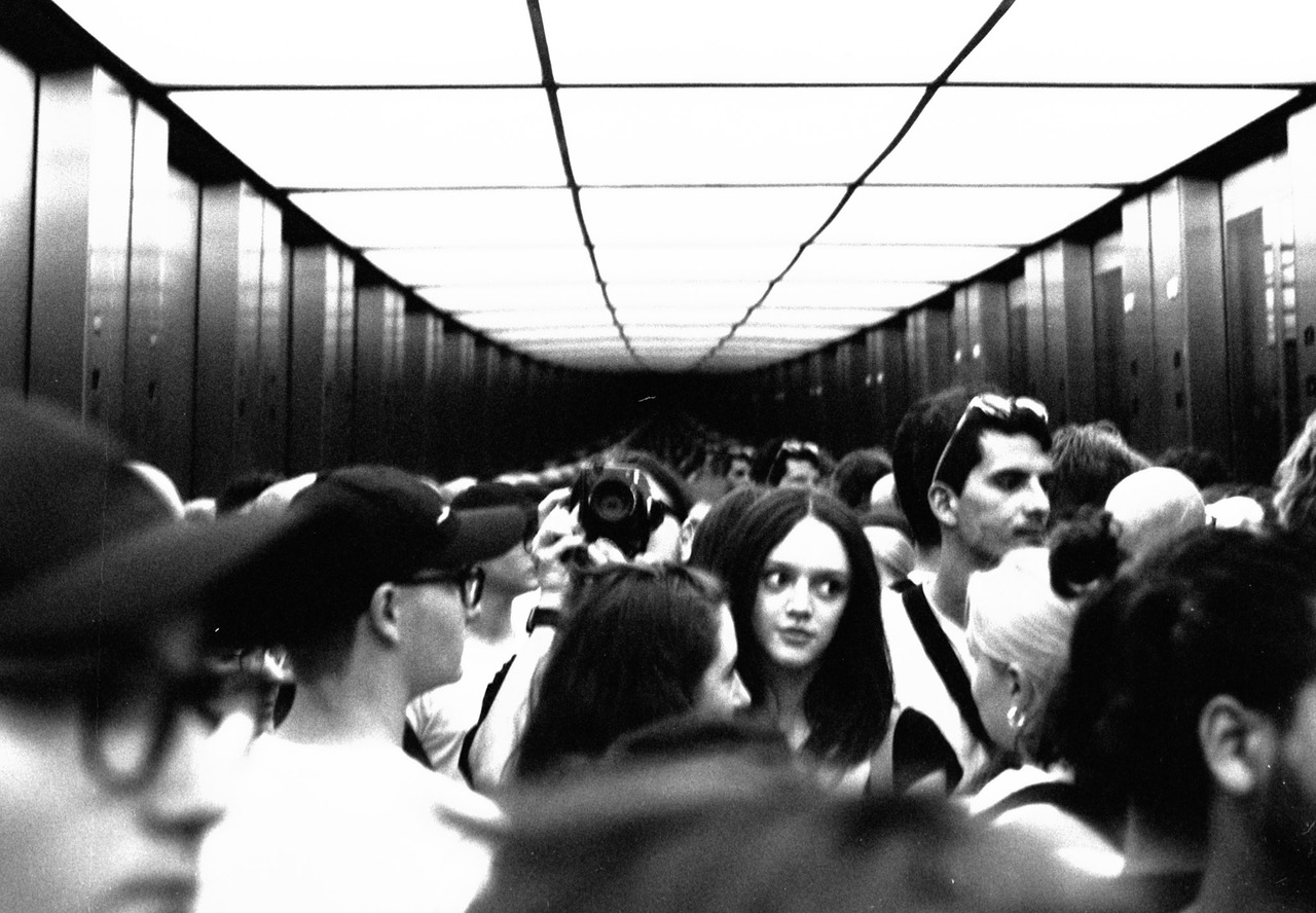 A crowded mirrored lift, a young woman in the centre looking toward the camera.